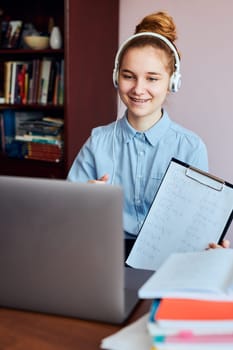 Young woman student showing homework, having classes, learning online, watching lesson remotely, listening to professor, talking with classmates on video call from home during quarantine. Young girl using laptop, headphones, books, manuals sitting at a desk