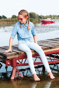 Young girl sitting on jetty over the lake and dipping feet in water on sunny day in the summertime