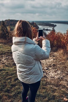 Back view of young woman taking photos of landscape with smartphone during trip on autumn sunny day