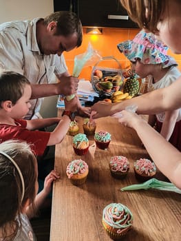 Group of children baking cupcakes, squeezing cream from confectionery bag, preparing ingredients, topping, sprinkles for decorating cookies. Kids cooking, working together in kitchen at home. Concept of happy family