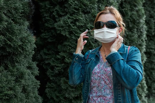 Woman walking outdoors wearing face mask during pandemic coronavirus outbreak standing at front of green tress