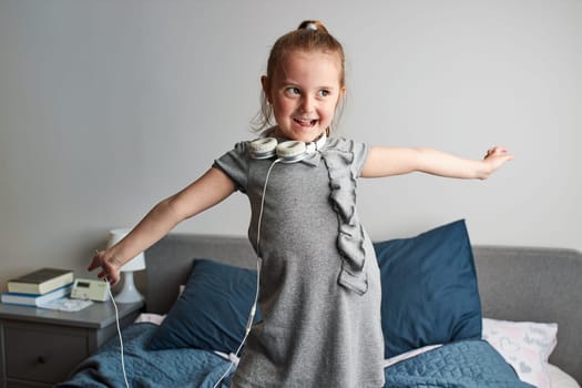 Little girl singing holding headphones cord imitating herself a real singer. Child having fun jumping dancing listening to music on bed in bedroom at home