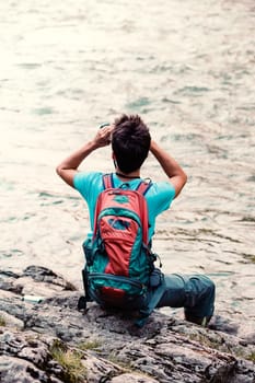 Young tourist with backpack looks through a binoculars, sits on a rock over a river. Boy spends a vacation on wandering with backpack, he is wearing sports summer clothes