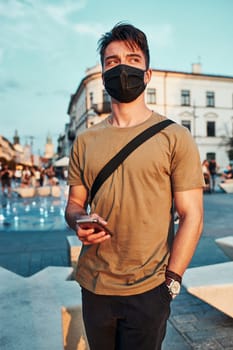Young man standing in the city center in the evening, looking away, wearing the face mask to avoid virus infection and to prevent the spread of disease in time of coronavirus