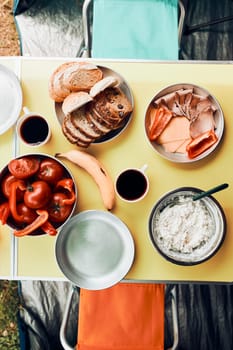 Breakfast prepared during summer vacation on camping. Bread, cottage cheese, cold meat, tomatoes, fruits and coffee cups on table. Close up of outdoor table setting set under tent