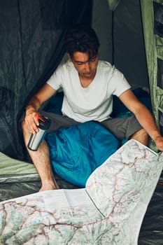 Young man looking at a map of mountain trails planning next trip on summer vacation sitting in a tent on camping. Concept of camp life
