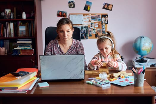 Woman mother working doing her job remotely during video chat call stream online course webinar on laptop from home while her daughter playing with bricks toy. Woman sitting at desk in front of computer looking at screen