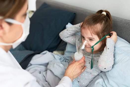 Doctor visiting little patient at home. Child having medical inhalation treatment with nebuliser. Girl with breathing mask on her face. Woman wearing uniform and face mask