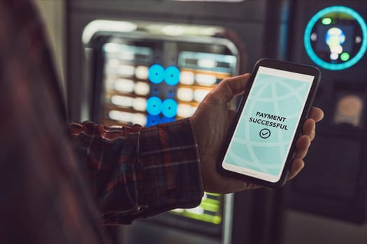 Consumer paying for product at vending machine using contactless method of payment with mobile phone. Man using payment app on smartphone to buy product. Male hand holding smart phone with payment confirmation on screen