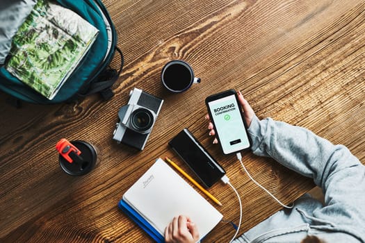 Woman planning next vacation trip. Booking hotel online using smartphone. Ordering service. Charging mobile phone with power bank. Using technology while travelling. Girl sitting at wooden table with camera and backpack during journey. Travel plan concept