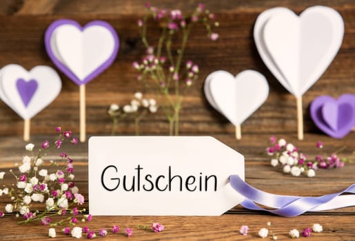 Label With German Text Gutschein Means Voucher. Purple And Lilac Decoration And Spring Flower Arrangement. Heart Symbols With Wooden Background.