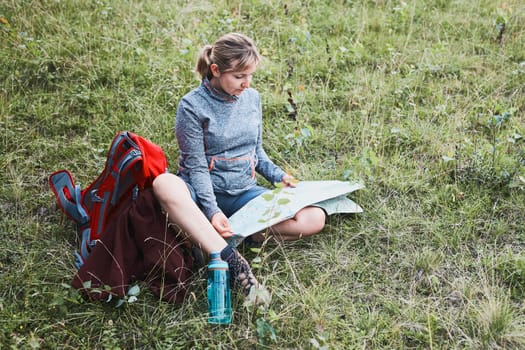 Woman with backpack having break during trip in mountains looking at map sitting on grass on summer vacation day