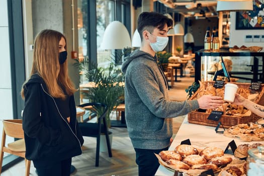 People standing in a queue to buy coffee and pastry in the coffee shop to go. Man taking coffee cup from barista. People standing at counter wearing the face masks to avoid virus infection and to prevent the spread of disease in time of coronavirus