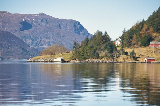 Fjord with mountains on horizon. Small cottages on the coast between stones, trees and meadow. Water glistens in the sun in Norway. Landscape photo from the north