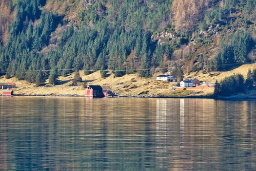 Fjord with mountains on horizon. Small cottages on the coast between stones, trees and meadow. Water glistens in the sun in Norway. Landscape photo from the north