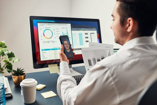 Man entrepreneur having business conversation, video chat on mobile phone. Businessman working with data, charts and graphs on computer screen. Man holding smartphone sitting at desk in office