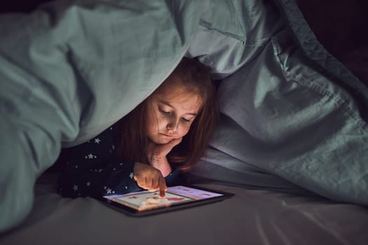 Little girl having fun watching, playing and listening to stories on tablet computer. Child having fun before bedtime laying under duvet using tablet