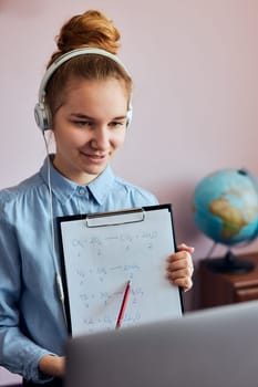 Young woman student showing homework, having classes, learning online, watching lesson remotely, listening to professor, talking with classmates on video call from home during quarantine. Young girl using laptop, headphones, books, manuals sitting at a desk