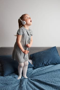 Little girl singing holding headphones cord imitating herself a real singer. Child having fun jumping dancing listening to music on bed in bedroom at home