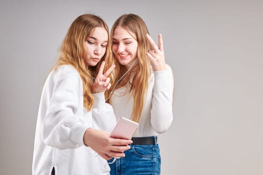 Young women girls listening to music together streaming content having fun watching video enjoying video chat talking with friends making gestures faces using smartphone earphones headphones standing over plain grey background