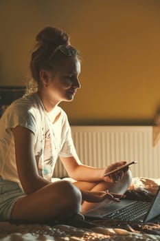 Young woman using portable computer and mobile phone, sitting on a bed, learning online at home. Candid people, real moments, authentic situations