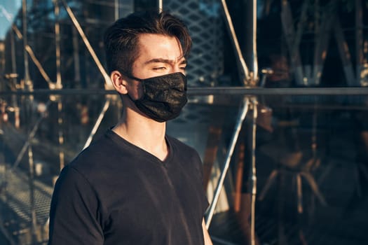 Young man walking in the city center along glass store front in the evening, looking away, wearing the face mask to avoid virus infection and to prevent the spread of disease in time of coronavirus