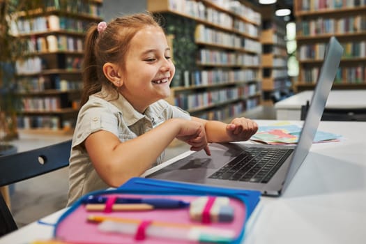 Cheerful student doing her homework using laptop in after school club at primary school. Back to school. Child using technology on elementary computer science class. Smart little girl learning from educational application at school. Child solving riddles and puzzles on personal computer