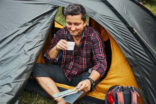 Man relaxing in tent at camping during summer vacation. Man holding map while planning next trip. Actively spending vacations outdoors close to nature. Concept of camp life