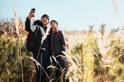 Hugging couple taking selfie while vacation trip. Hikers with backpacks on way to mountains. People walking through tall grass along path in meadow on sunny day. Active leisure time close to nature