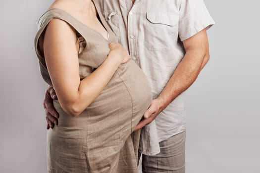 Pregnant woman and her husband touching her tummy standing over plain background. Pregnancy, expectation, motherhood concept