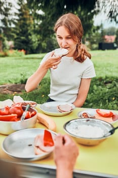 Family having breakfast outdoors on camping during summer vacation. Bread, cottage cheese, cold meat, tomatoes, fruits and coffee cups on table. Close up of outdoor table setting set on grass in the garden