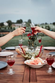 Couple making toast during summer outdoor dinner in a home garden. Hands holding wine glasses with red wine over table with dishes