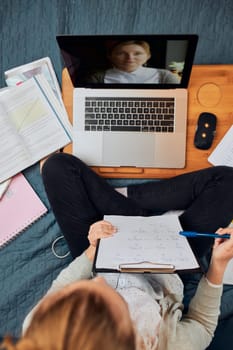 Young woman student having classes, learning online, watching lesson remotely, listening to professor, talking with classmates on video call from home during quarantine. Young girl using laptop, headphones, books, manuals sitting on bed