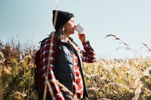 Woman taking break and enjoying the coffee during vacation trip. Woman standing on trail and looking away holding cup of coffee. Woman with backpack hiking through tall grass along path in mountains. Spending summer vacation close to nature