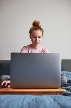 Teenage girl working on school tasks homework remotely on her laptop from home during COVID-19 quarantine. Online course lessons. Learning remotely. Girl sitting on bed in front of computer looking at screen
