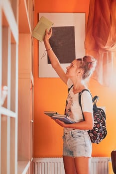 Teenage girl looking for books in local library. Young woman with a backpack standing by a bookshelf indoors. Real people, authentic situations