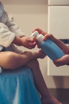 Mother applying moisturizing cream on her daughter's legs after bath. Mom caring about her child. Girl sitting in bathroom, wearing bathrobe