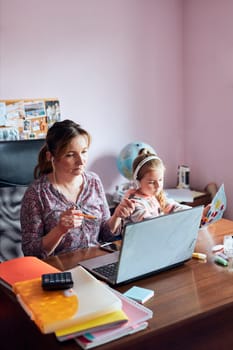 Woman mother working doing her job remotely during video chat call stream online course webinar on laptop from home while her daughter making drawings playing around and listening to stories. Woman sitting at desk in front of computer looking at screen