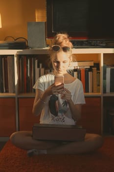 Young woman using portable computer and mobile phone, sitting on a floor, learning online at home. Candid people, real moments, authentic situations