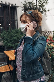 Young woman talking on phone standing in a street downtown. Woman is wearing the face mask to avoid virus infection and to prevent the spread of disease in time of coronavirus