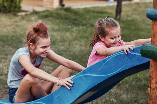 Teenage girl playing with her younger sister in a home playground in a backyard. Happy smiling sisters having fun on a slide together on summer day. Real people, authentic situations