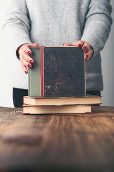 Young woman holding a few old books on wooden table in antique bookstore. Woman wearing grey sweater and jeans. Vertical photo