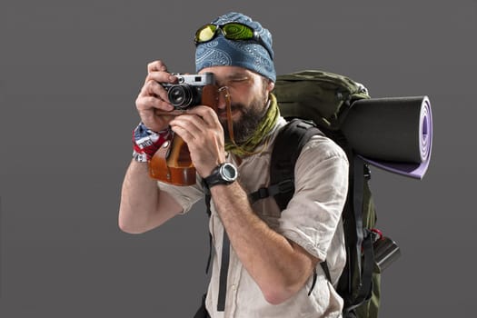 The tourist with camera. Portrait of a male fully equipped tourist with backpack. He photographing something on gray background