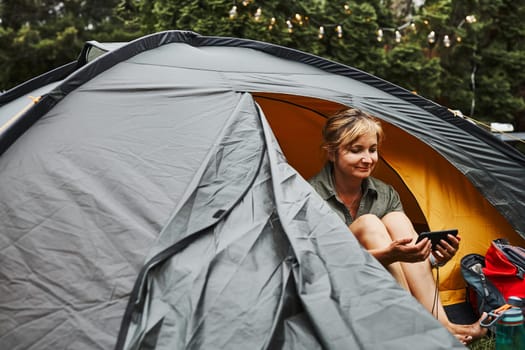Woman relaxing in tent at camping during summer vacation. Female holding smartphone while video call with friends. Actively spending vacations outdoors close to nature. Concept of camp life