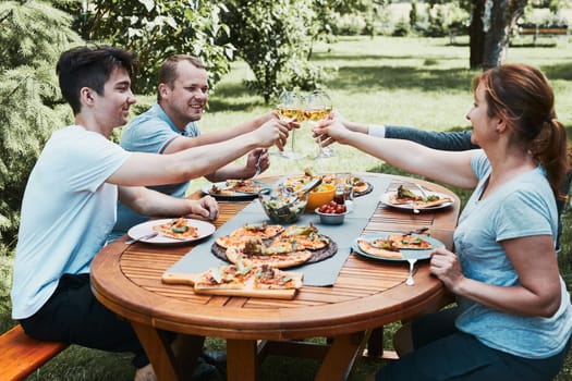 Friends making toast during summer picnic outdoor dinner in a home garden. Close up of people holding wine glasses with white wine over table with pizza, salads and fruits. Dinner in a orchard in a backyard
