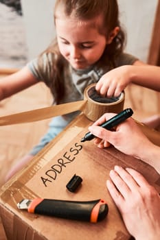 Woman writing an address on a cardboard box parcel in room at home. Little girl helping her mother to wrap a package. Preparing parcel to send