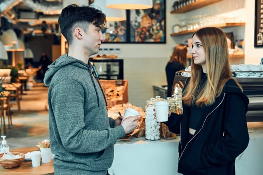 Friends having a chat, holding coffee cups, standing in a coffee shop and waiting for the orders. Man and woman having a break drinking coffee in cafe