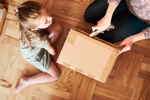 Woman unpacking a cardboard box parcel in room at home. Little girl waiting for opening a gift in package