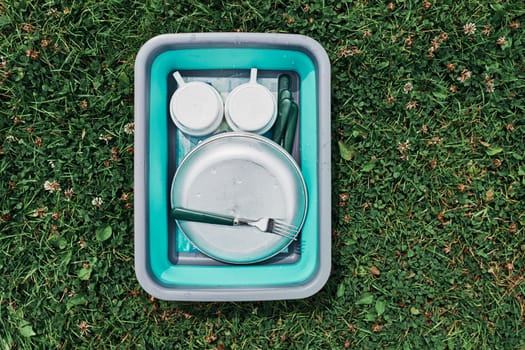 Washing up bowl filled with the washed outdoor dishes, plates, cups and cutlery put on grass on camping