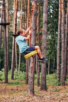 Happy boy riding on the zip line in rope park in forest while spending summer vacation. Real people, authentic situations